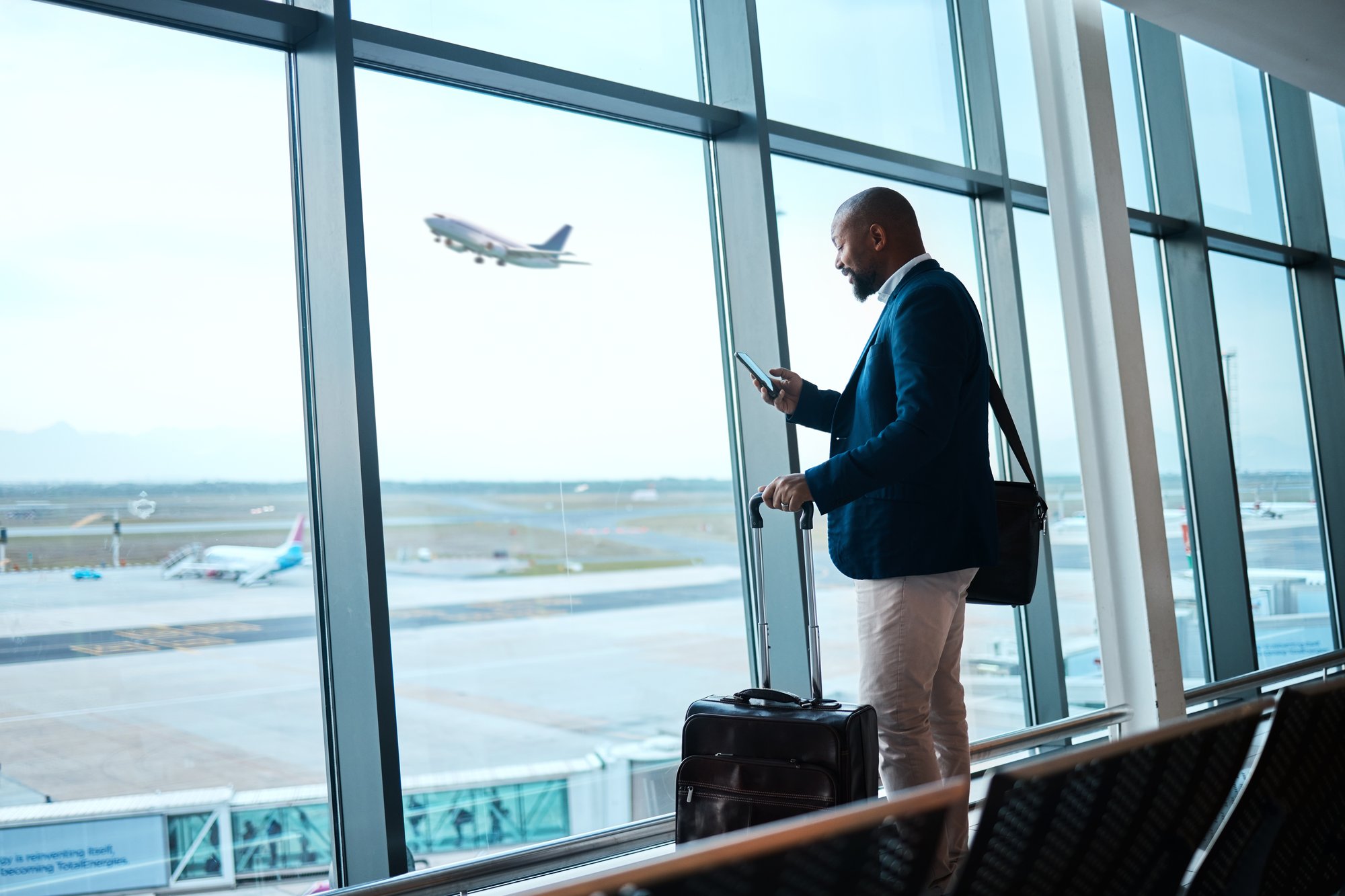 Business traveler in an airport terminal checking a phone, suitcase beside them, airplane visible outside the window.
