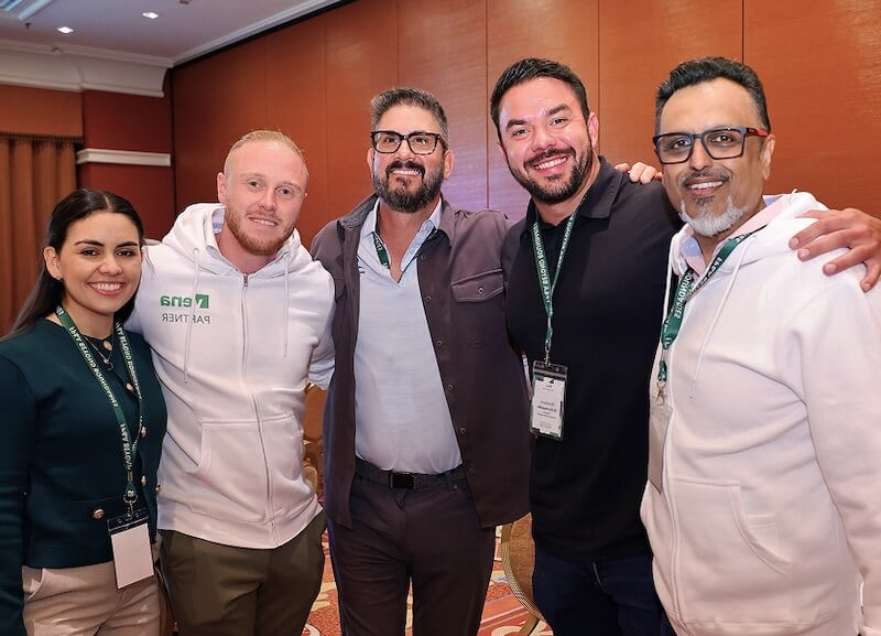 Five people wearing conference lanyards smile and pose together in a hotel meeting room.