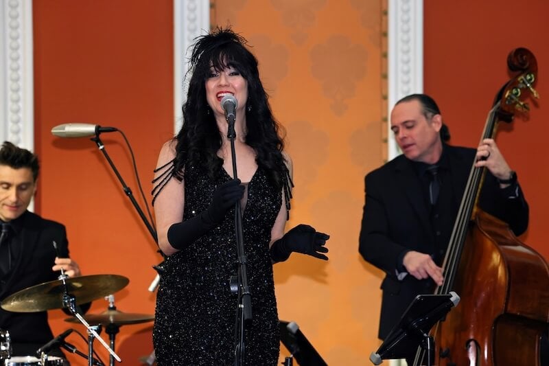 Female singer in a black sequined dress and gloves singing into a microphone with drummer and upright bassist behind her