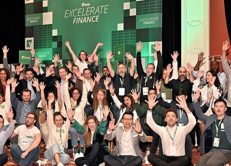 Large group of conference attendees seated and standing with hands raised in front of a green 
