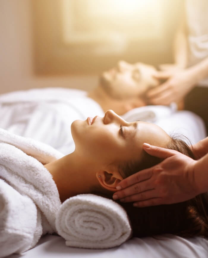 Woman lying on a rolled towel receiving a gentle head massage, with a man receiving a similar treatment in background.