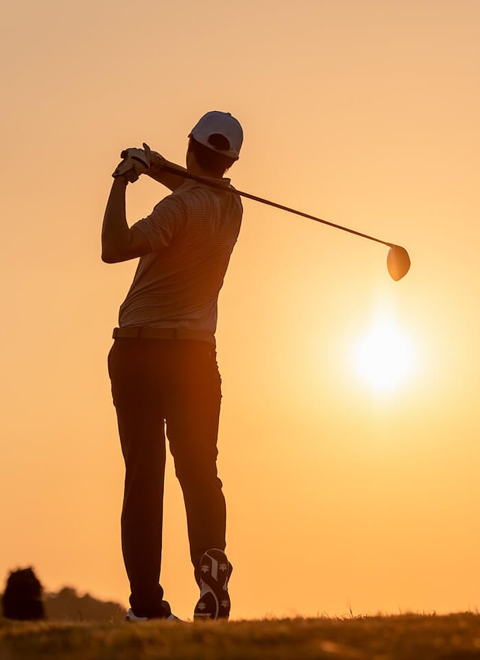 Golfer finishing a swing with a driver, silhouetted against an orange sunset.