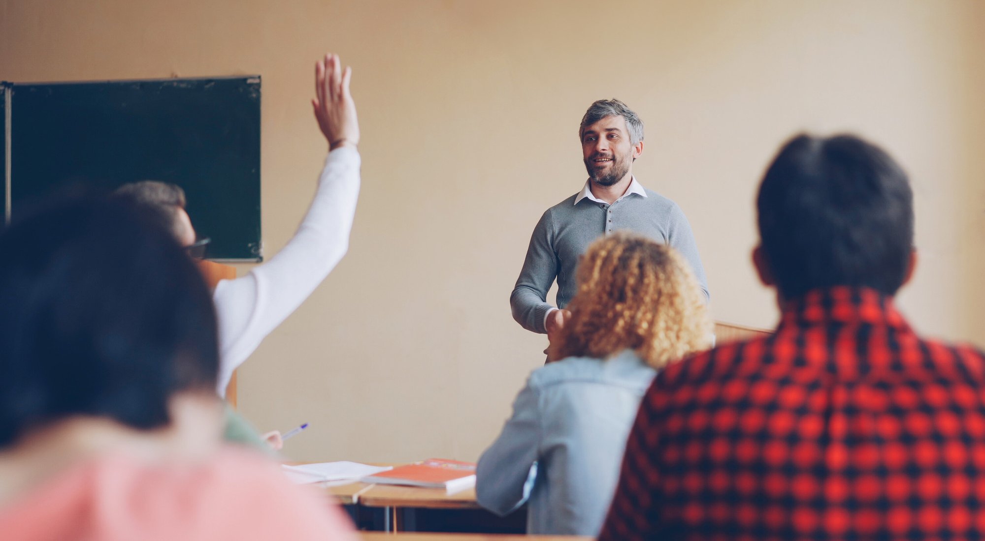Friendly male teacher speaking to his students, young man is raising hand and answering question