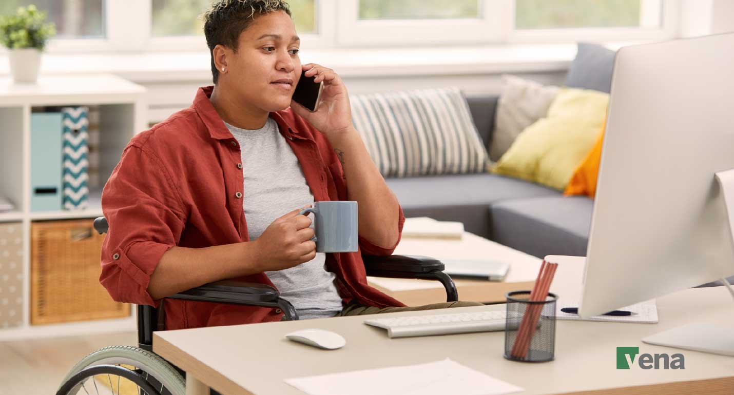 A wheelchair user at a desk working from the comfort of their home, talking on a cell phone and holding a cup of coffee