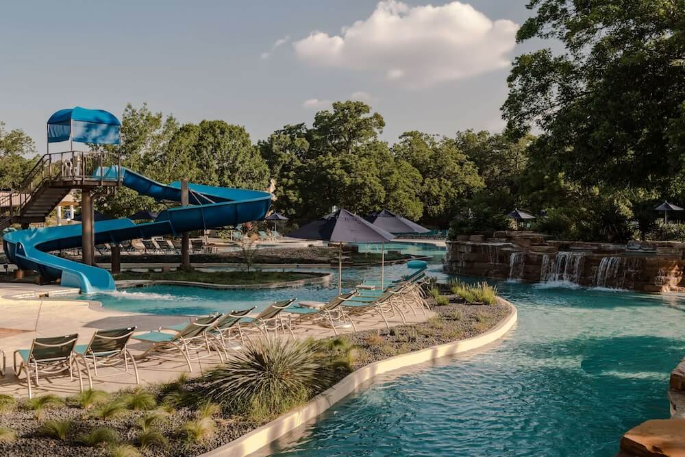Resort pool with winding blue water slide, lounge chairs, umbrellas, and small waterfalls surrounded by trees