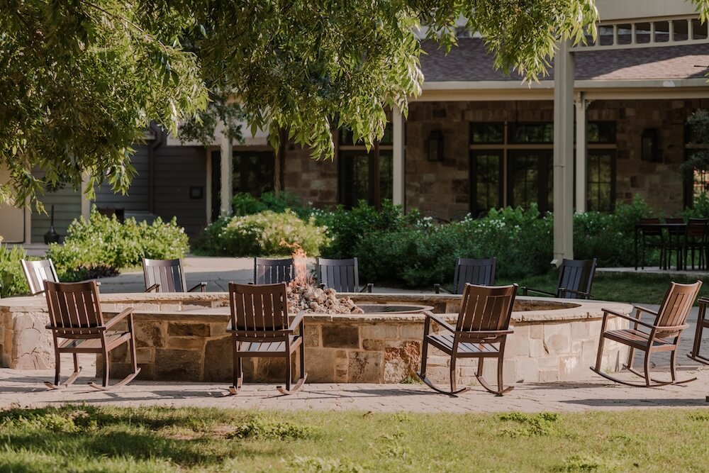 Outdoor stone fire pit with small flame, surrounded by empty wooden chairs in a garden courtyard.