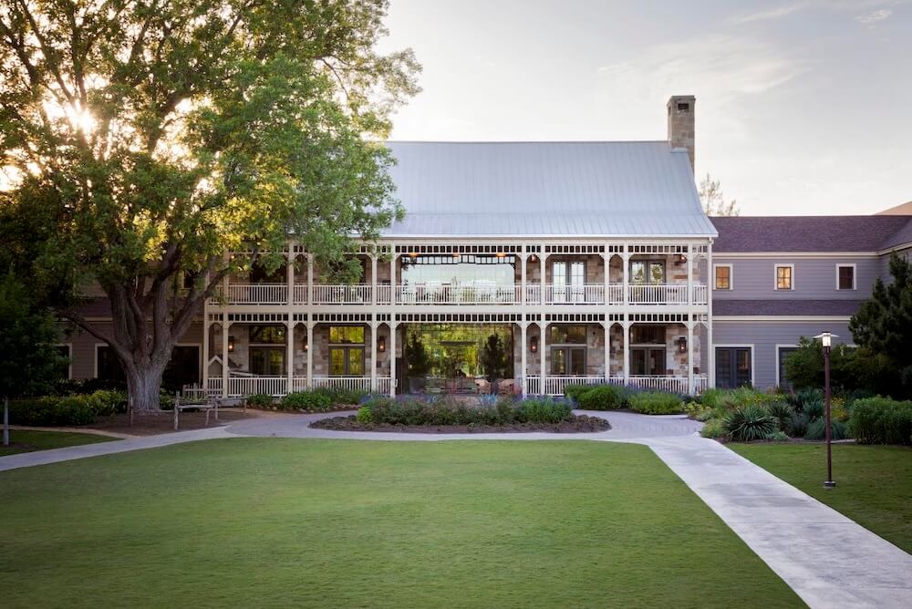 Large farmhouse-style building with two-story wraparound porches, big tree, and manicured lawn at sunset.