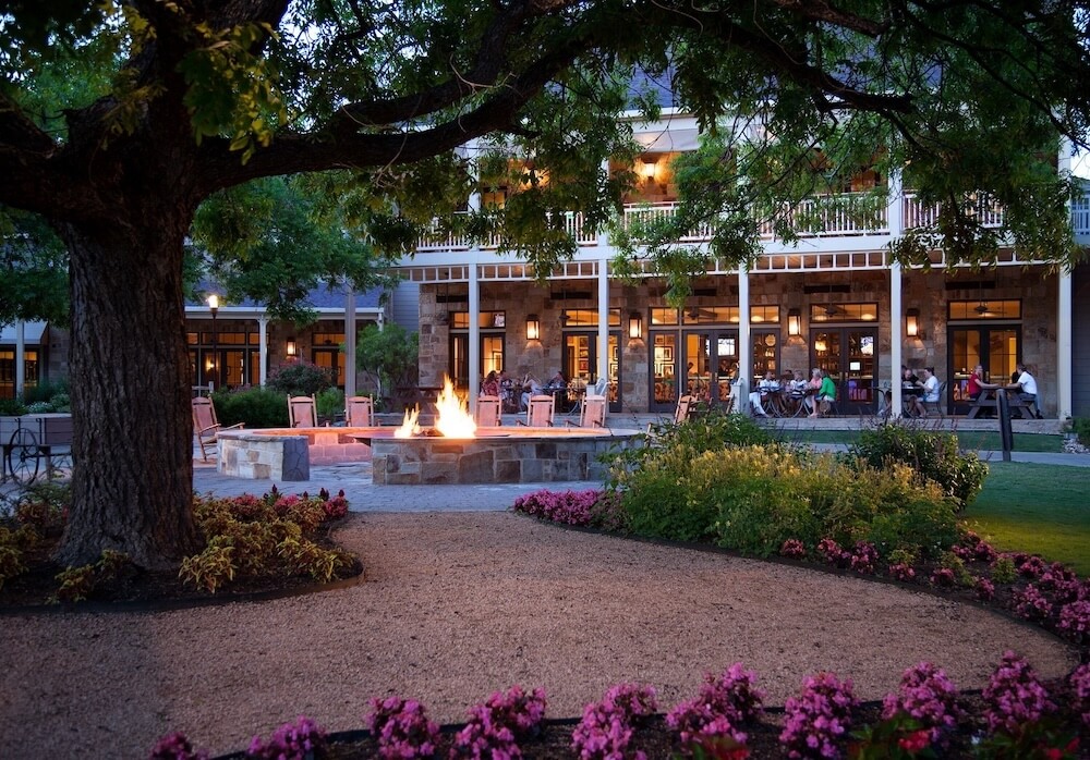 Courtyard at dusk with a central stone fire pit, surrounding chairs, flowering beds, and a two-story lit building.