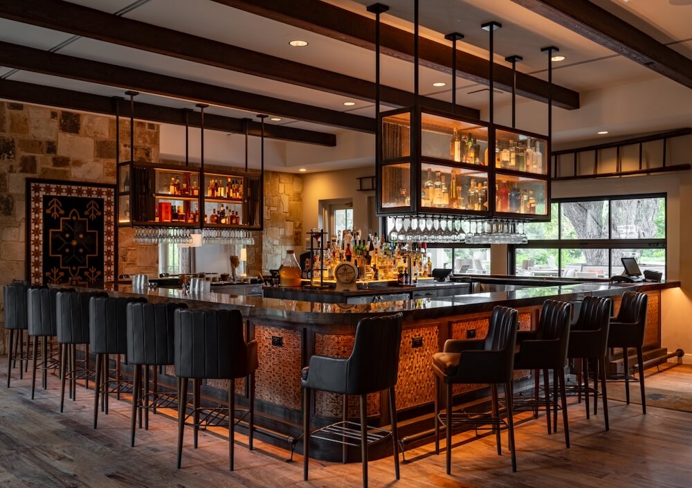 U-shaped bar with black stools, hanging bottle shelves, and warm lighting in a rustic room with stone walls and wood beams.