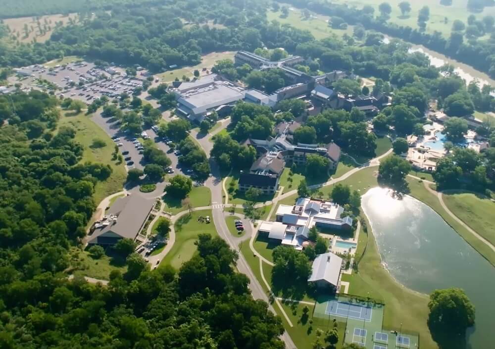 Aerial view of Lost Pines Resort with parking lots, winding roads, tennis courts, a pond, and surrounding trees.
