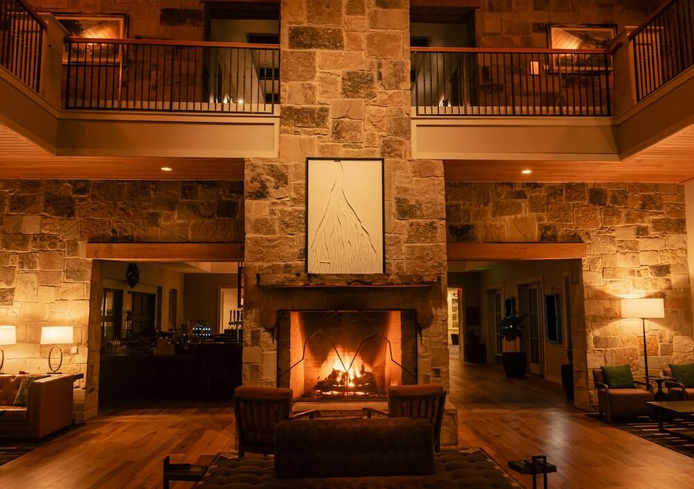 Cozy lodge interior with a stone fireplace burning, warm lighting, and seating arranged around the hearth.