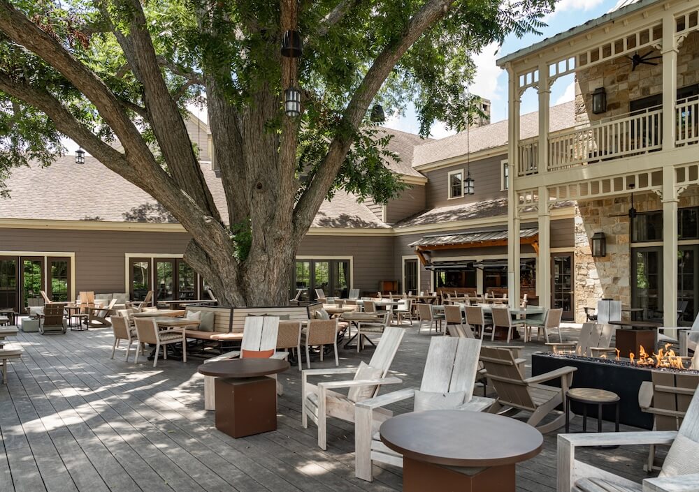 Spacious outdoor patio with wooden seating, fire pits, and a large shade tree beside a rustic lodge-style building.