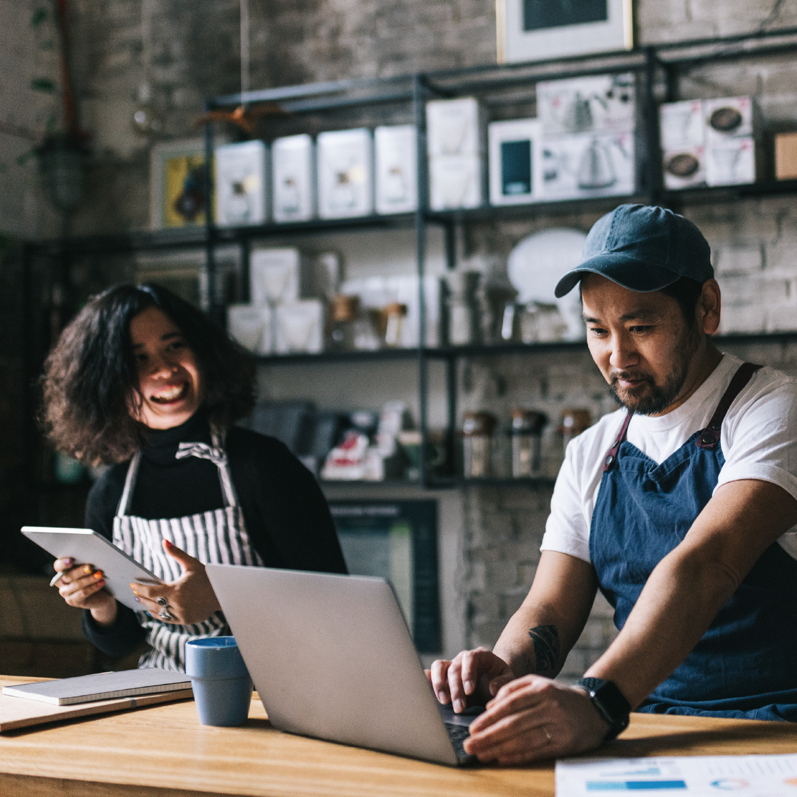 Two coffee shop employees standing behind a counter, one holding a tablet and one using a laptop