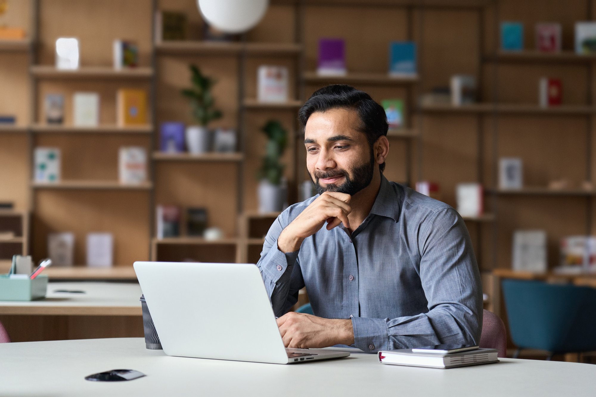 Smiling bearded indian businessman working on laptop in modern office lobby space