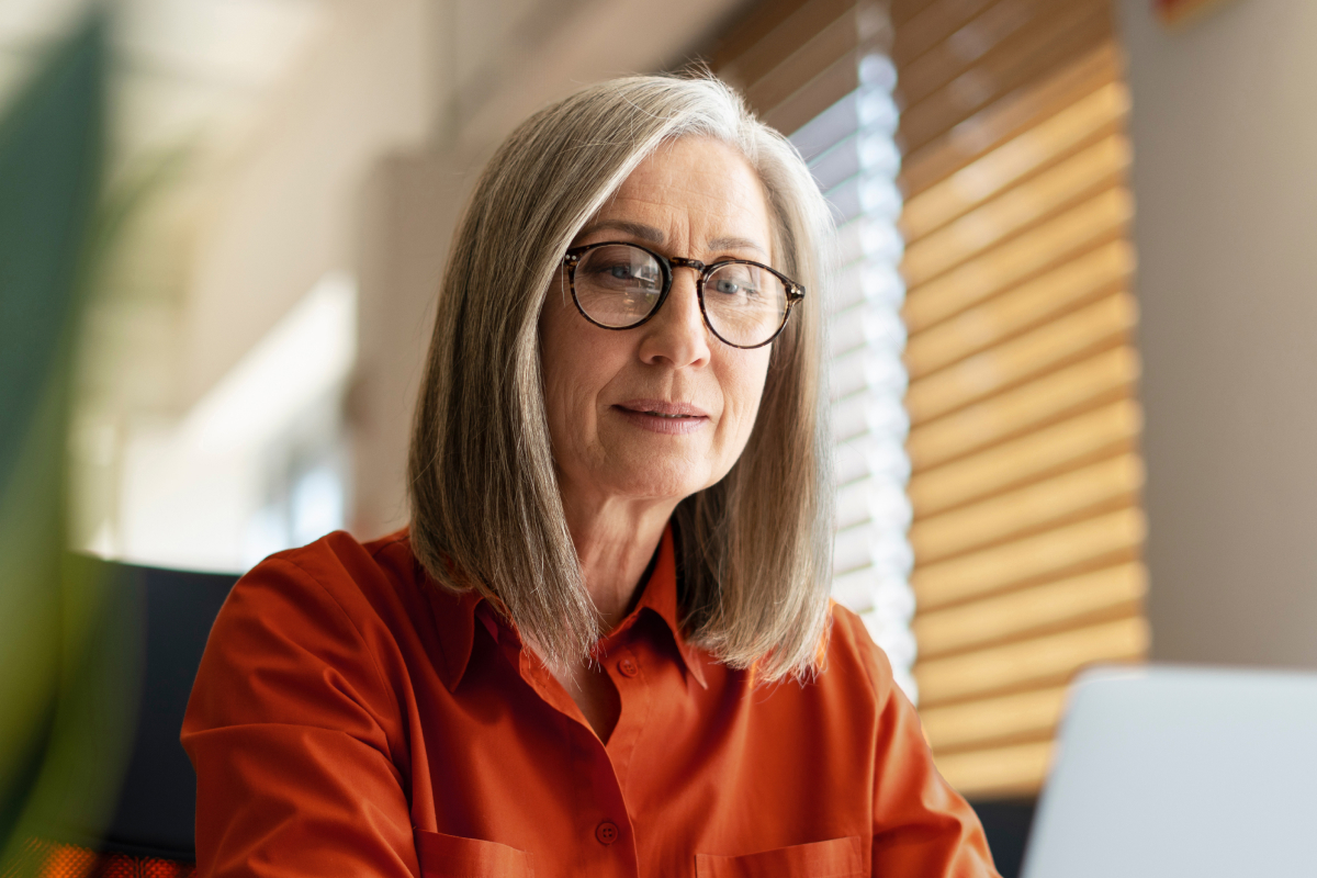 A woman wearing glasses sits at a desk, focused on her laptop while working in a well-lit office environment.