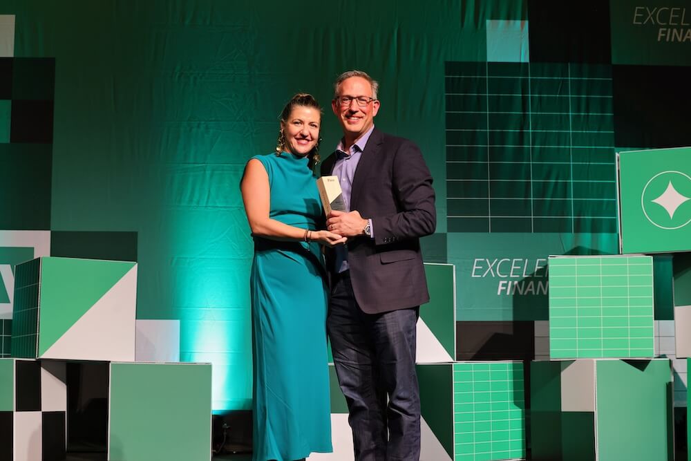 Two people onstage smiling and holding an award amid green geometric backdrops