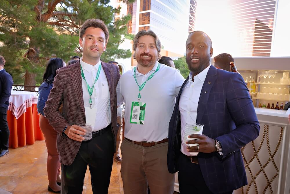 Three men with event lanyards smile at an outdoor reception, holding drinks near a bar with bottles and glassware.