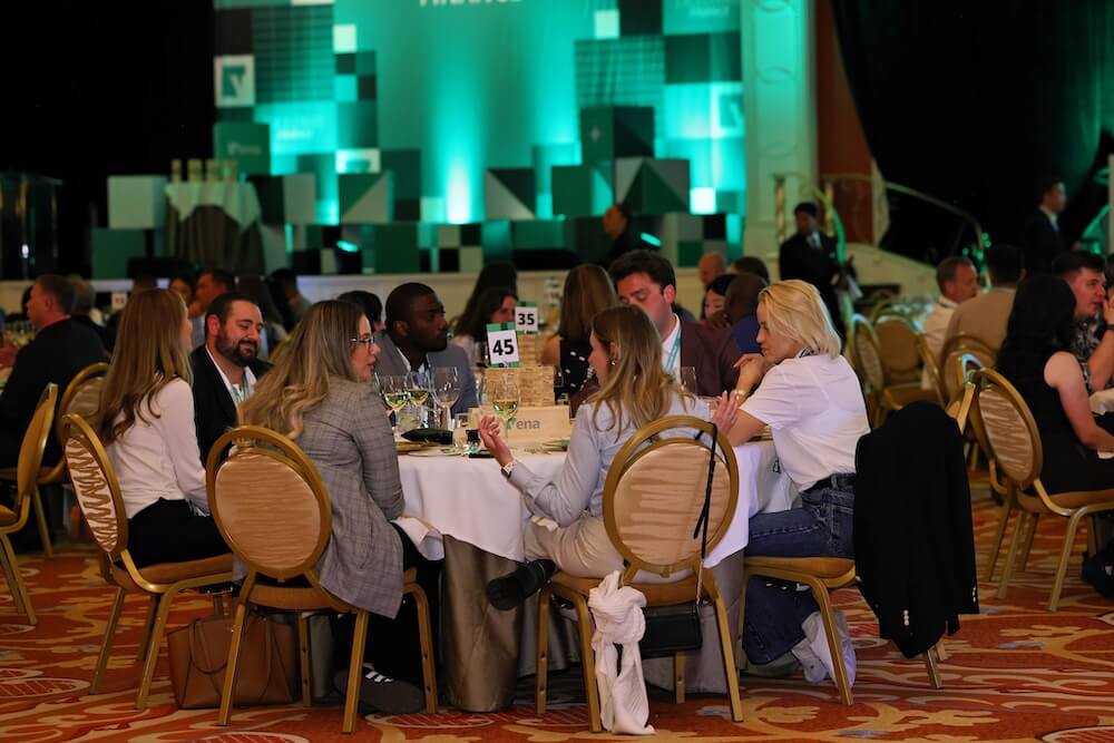 People seated at round tables chatting during a formal banquet with a green-lit stage in the background.