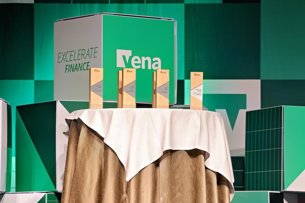 Four wooden awards on a draped round table with “Vena” and “Excelerate Finance” branding in a green stage backdrop.