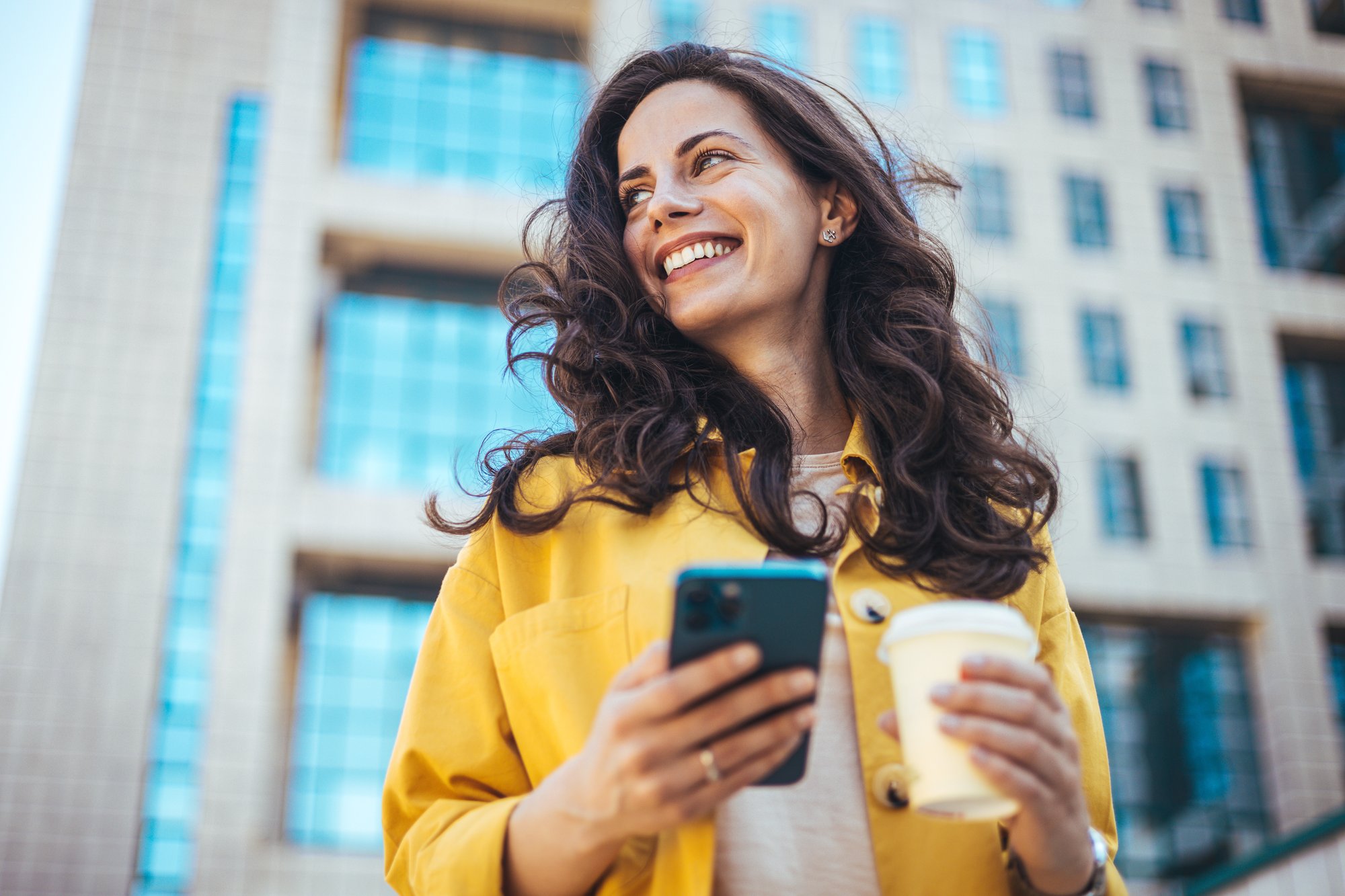Young positive businesswoman with take away coffee cup in hand using mobile phone while standing on sunny street near office building