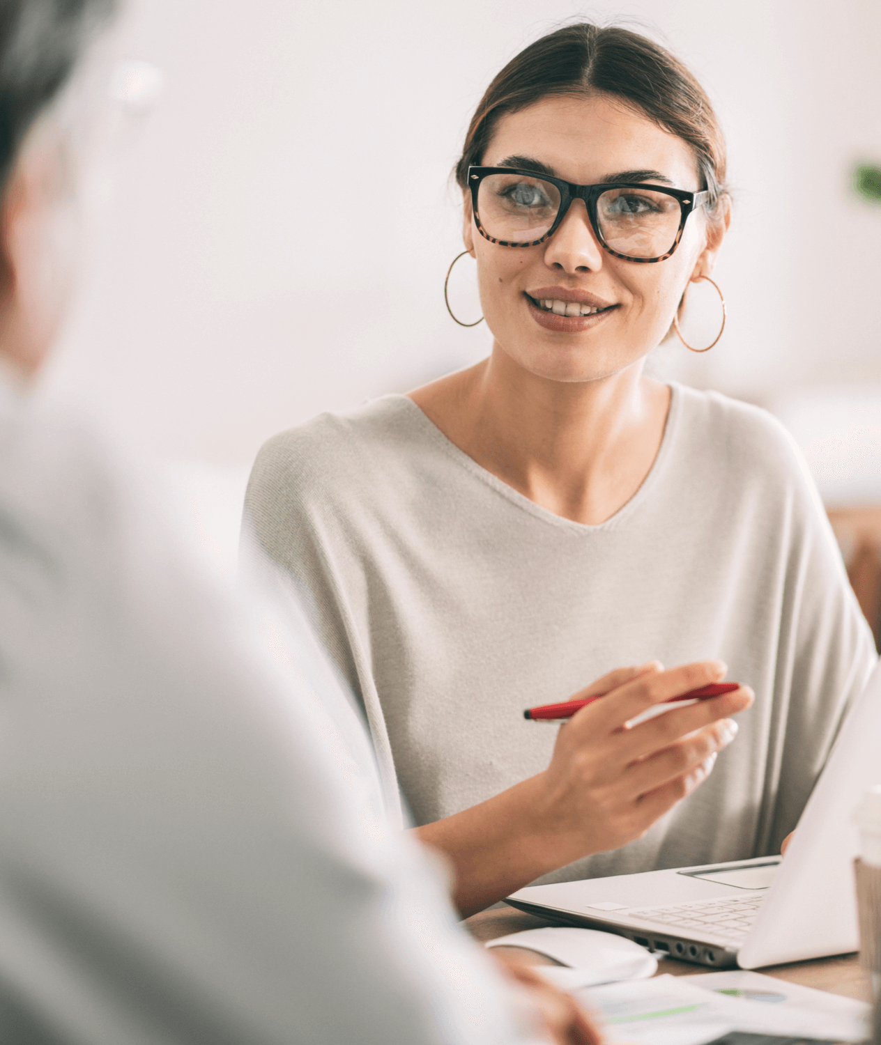 Caucasian woman with brown hair and glasses speaking with a man at a desk.