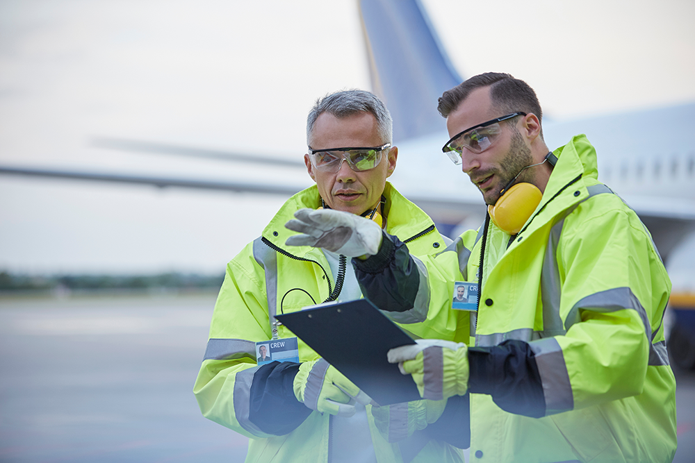 Two airport ground crew in high-visibility jackets reviewing a clipboard on the tarmac near an aircraft.