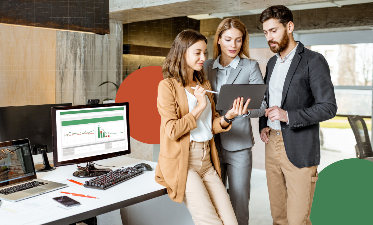 Two white women and one white man stand in an office setting, with one of the women showing financial reports on a tablet to her two colleagues 