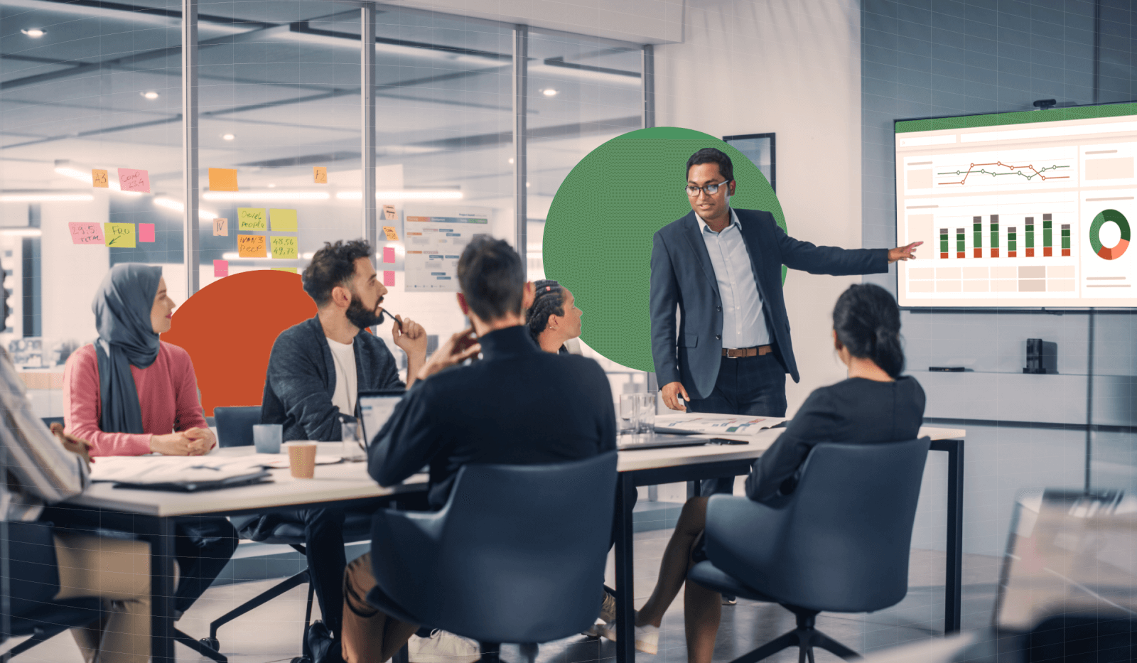 A man is presenting graphs on a screen at the front of the room while six other business professionals sit at a table engaged with the presenter. 